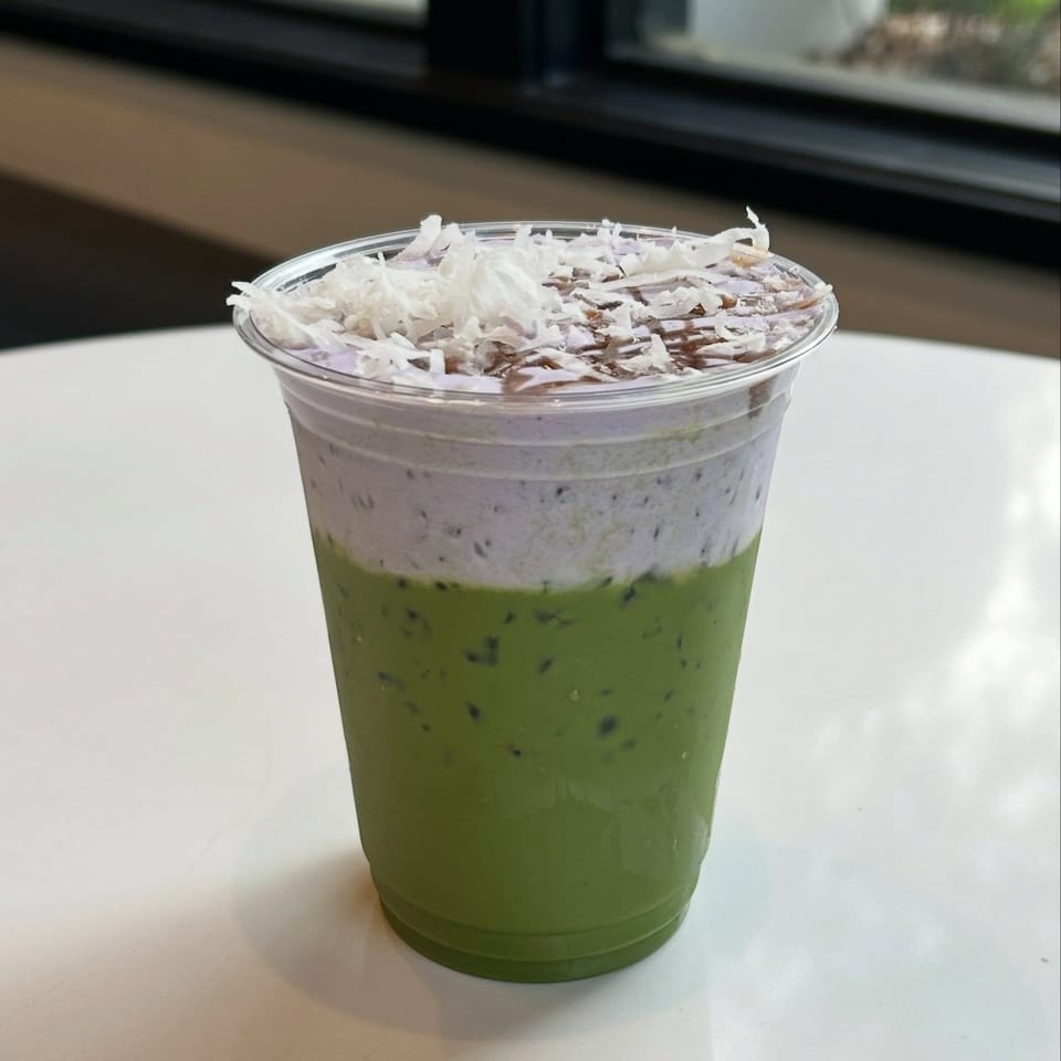 Green iced drink topped with whipped cream, shredded coconut, and chocolate shavings in a clear plastic cup on a table.