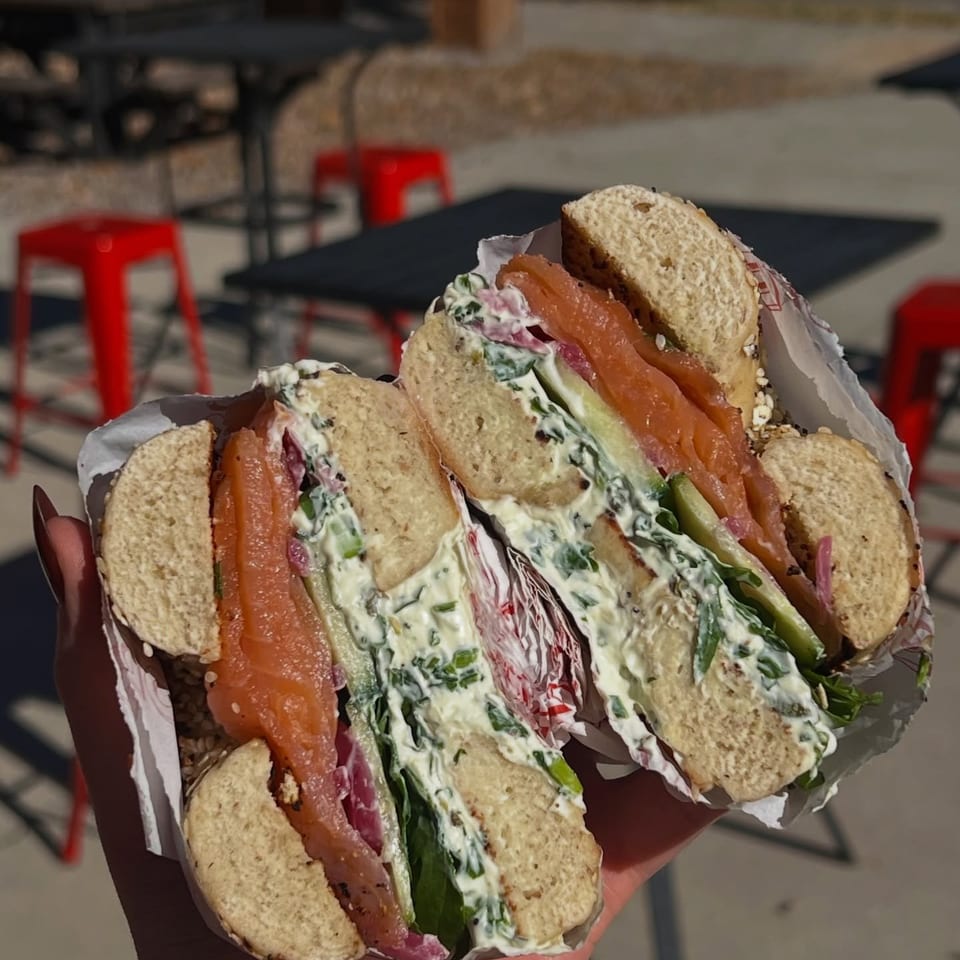 Handheld sesame bagel sandwich filled with smoked salmon, cucumber, greens, and creamy herb spread, with red stools outdoors.