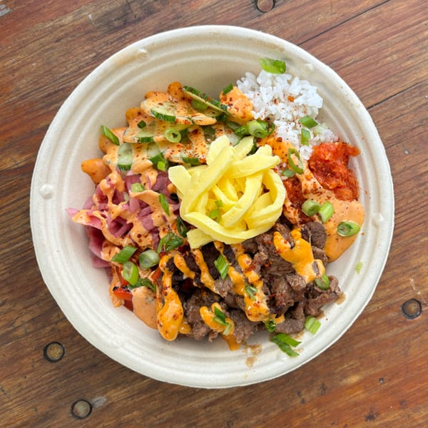 Colorful Korean BBQ bowl with beef, rice, kimchi, pickled vegetables, and sauces on a wooden table.