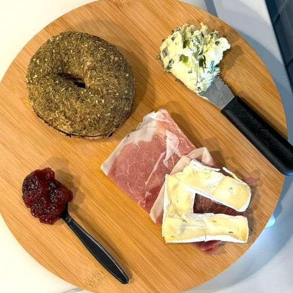 Savory bagel, prosciutto, brie, jam, and herb cream cheese on a round wooden board.