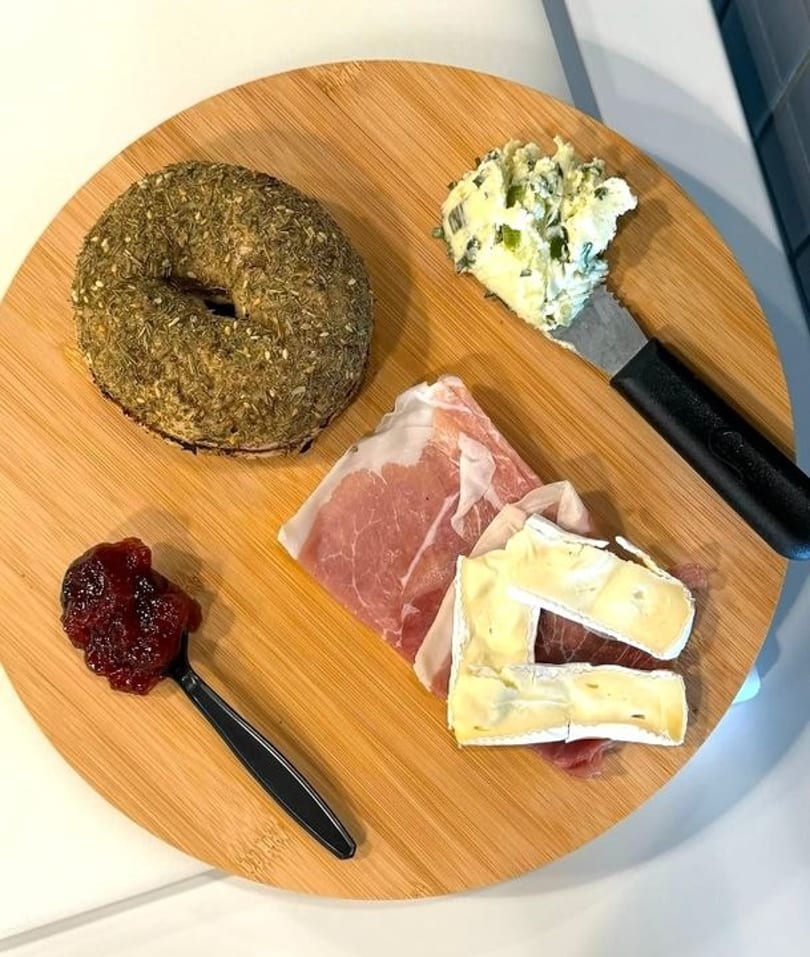 Savory bagel, prosciutto, brie, jam, and herb cream cheese on a round wooden board.