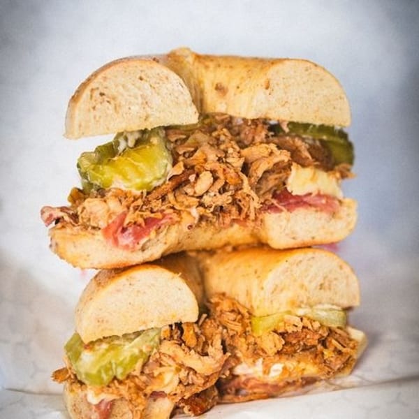 Two Cuban sandwiches on bagels, stacked, filled with pulled pork, ham, cheese, and pickles.