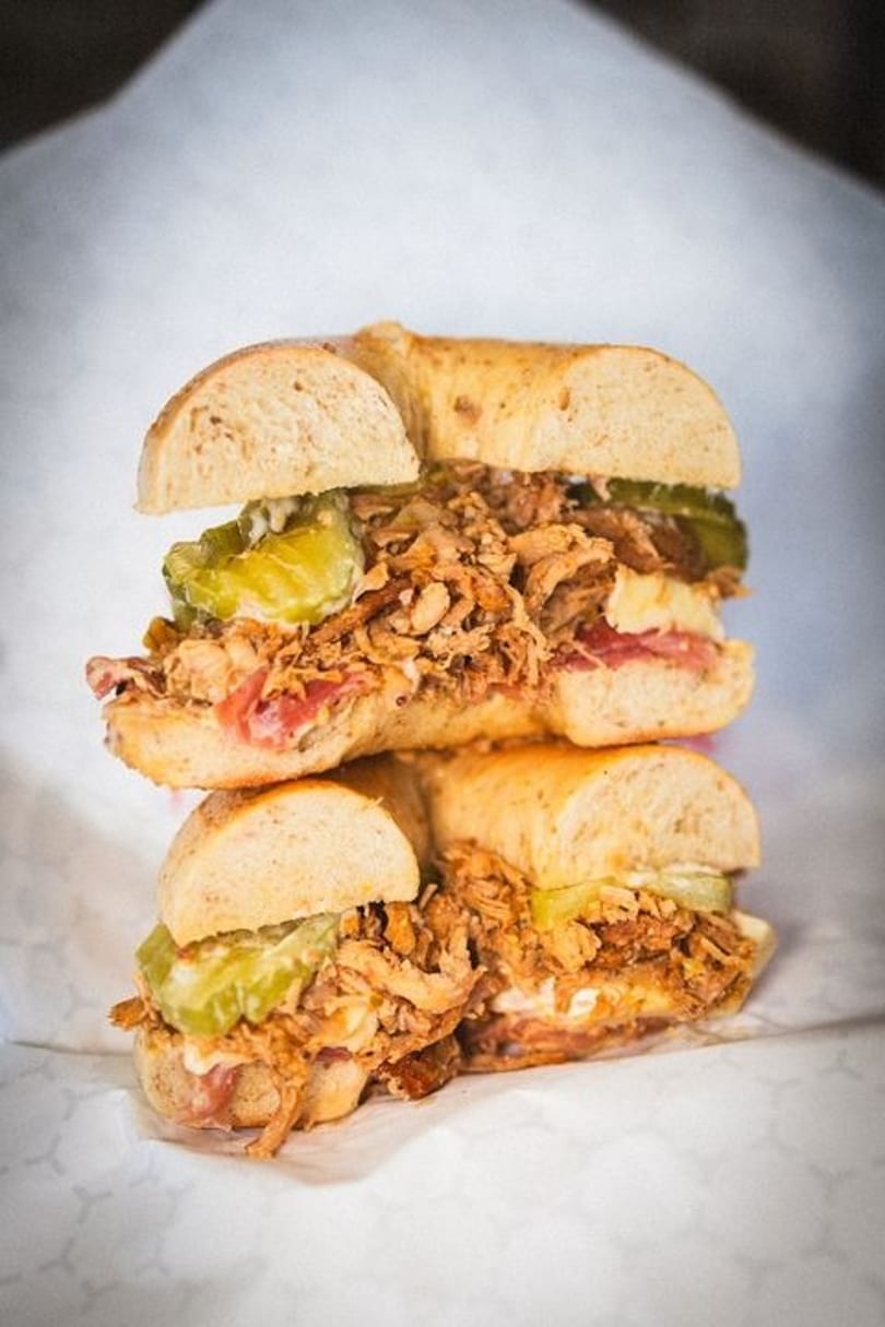 Two Cuban sandwiches on bagels, stacked, filled with pulled pork, ham, cheese, and pickles.