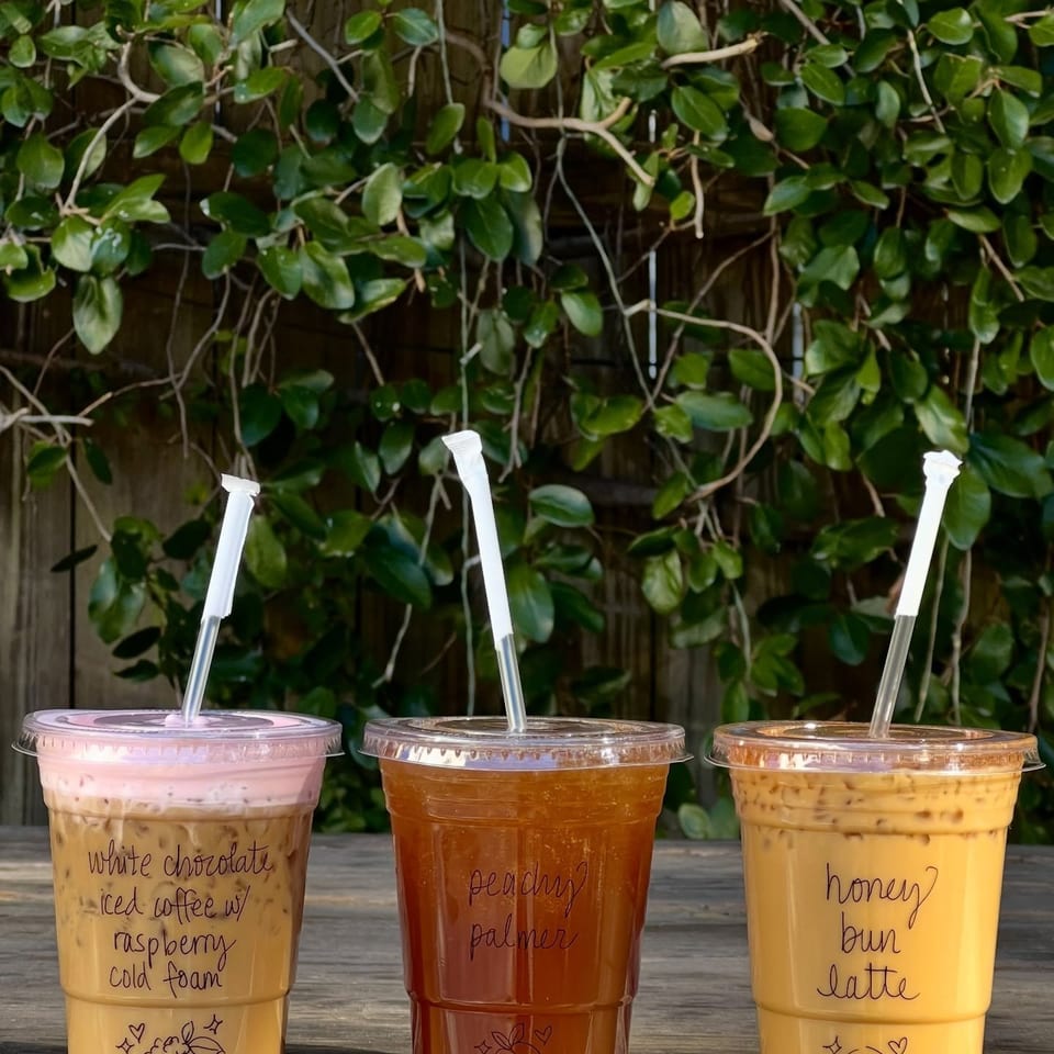 Three colorful iced drinks with handwritten labels sit on a wooden table against a leafy vine wall.