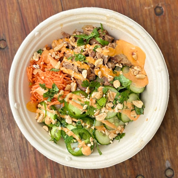 Close-up of a colorful bowl with meat, shredded carrots, sliced cucumbers, and peanuts, drizzled with sauce.
