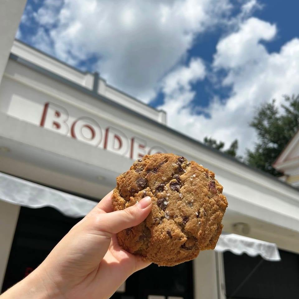 Hand holding a large chocolate chip cookie outside a bakery beneath a bright, cloud-filled sky.